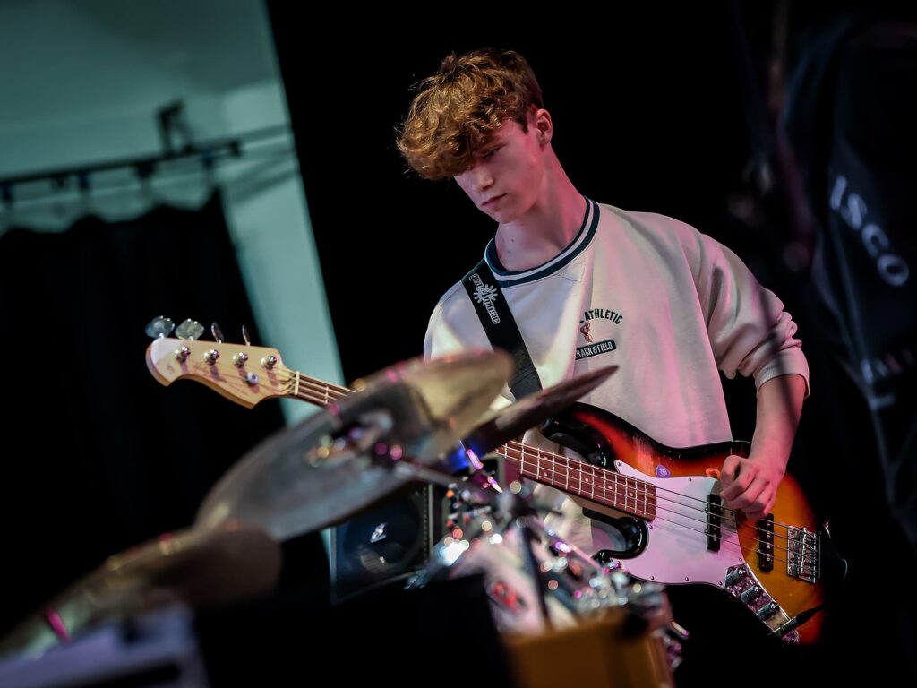 A young male playing the bass guitar in a band. He is focused and driven