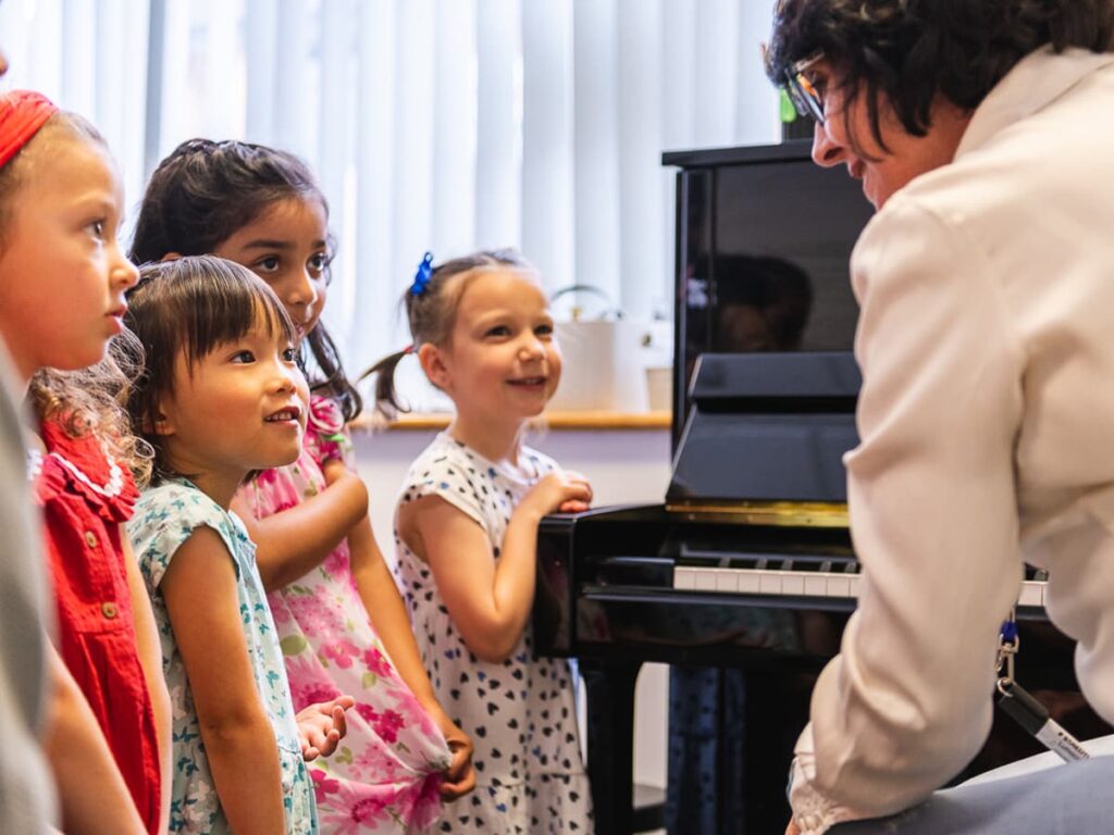 A group of young children attentively listening to a music teacher near a piano in a bright classroom.