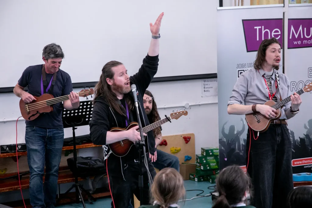 A group of musicians performing with ukuleles, a bass guitar, and percussion during a school music workshop by The Music Works.