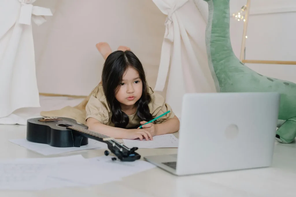 A young girl lying on the floor with a ukulele, taking notes while watching an online music lesson on her laptop.