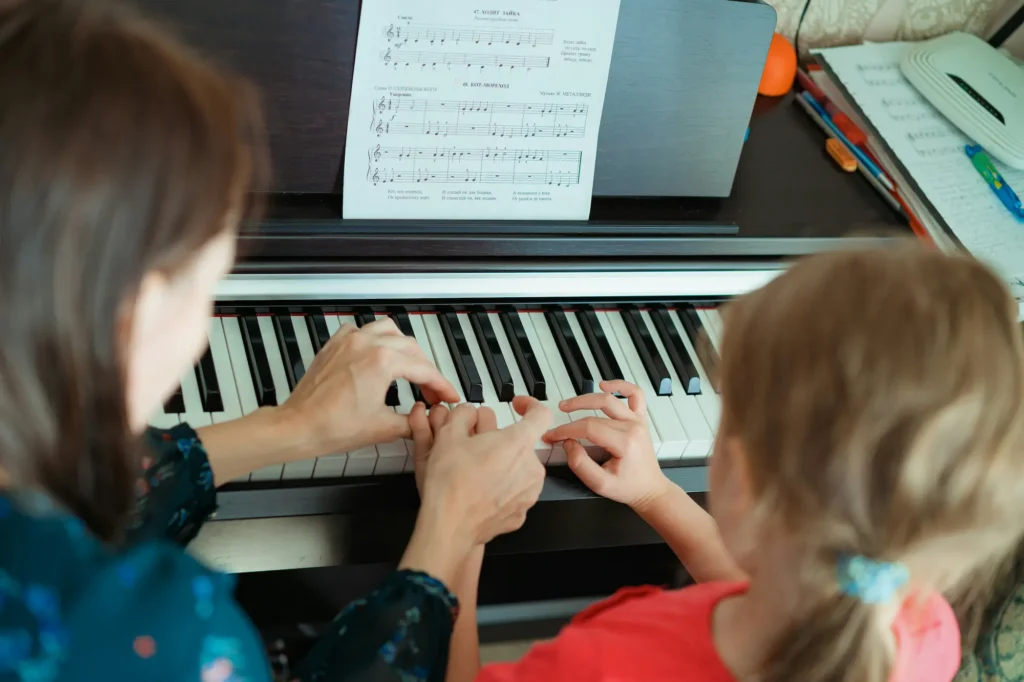 A child learning to play piano with a teacher guiding her hands on the keyboard.