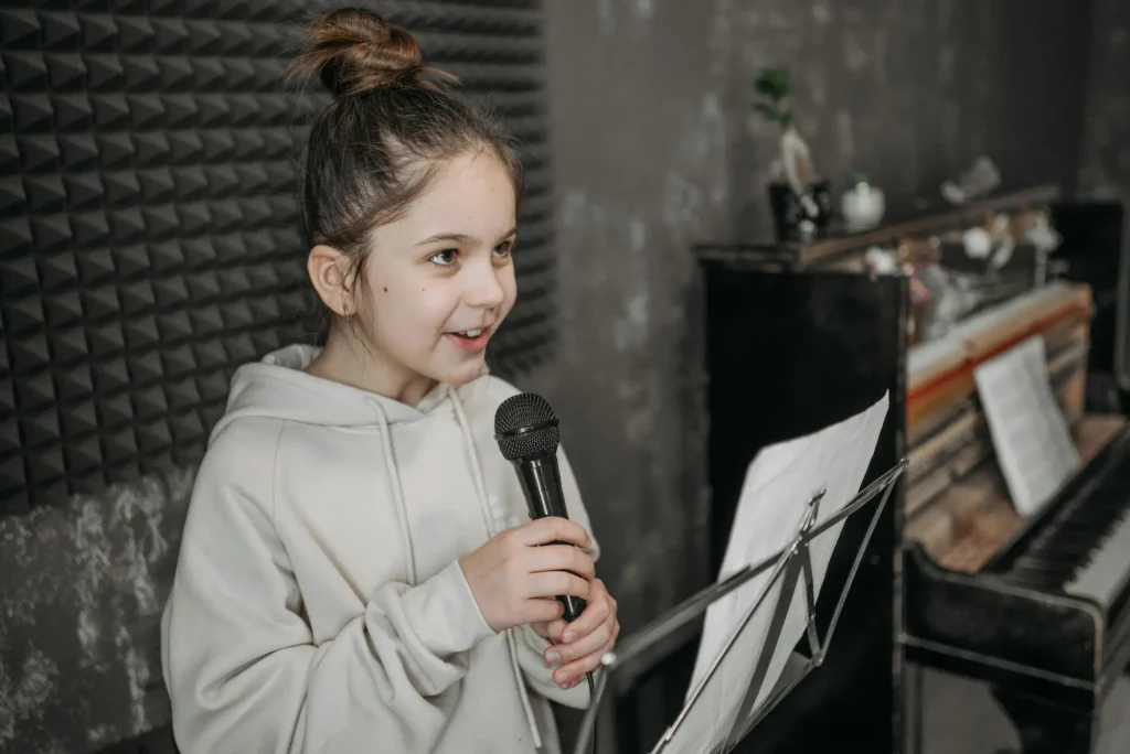 A young girl in a recording studio holding a microphone and singing from a music stand.