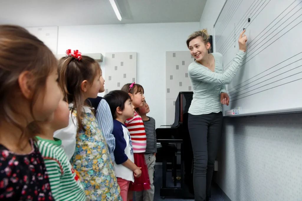 A group of young children attentively watching their music teacher explain notes on a whiteboard in a classroom.