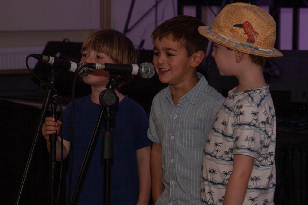 Three young boys singing into microphones during a live music performance at The Music Works.