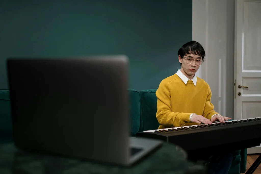 A young man in a yellow sweater playing a digital piano while attending an online music lesson on his laptop