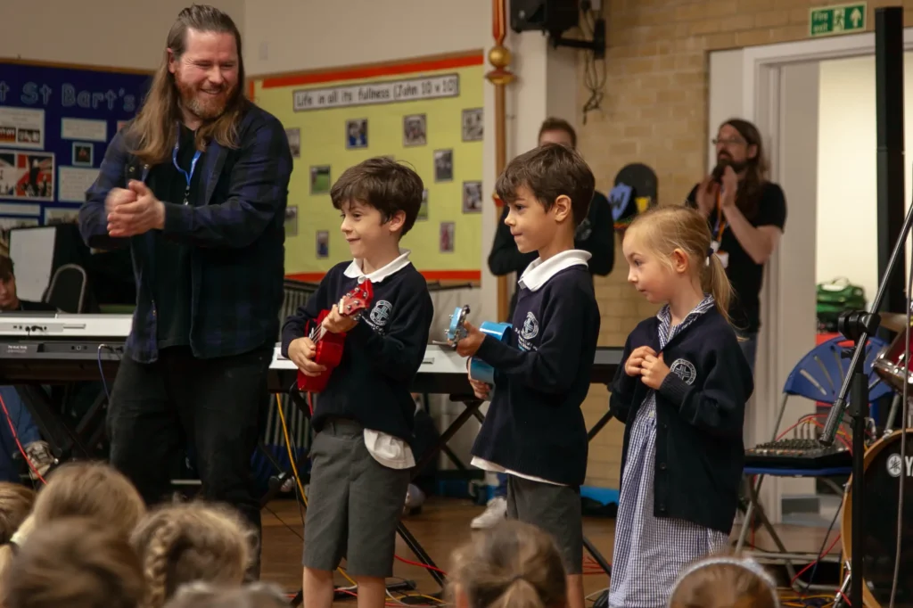 Music tutor from The Music Works applauding primary school children playing ukuleles in a school assembly.