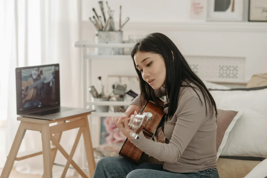 A young woman playing the guitar while following an online music lesson on her laptop.