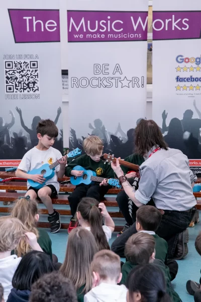 A music instructor from The Music Works teaching two young students to play the guitar during a school workshop.