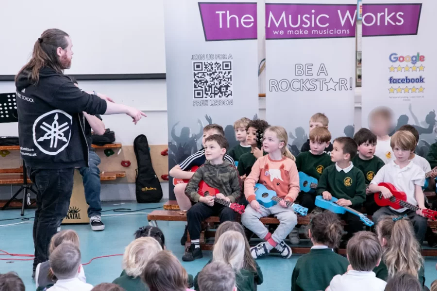 A music instructor from The Music Works guiding young students playing guitars during a school workshop.