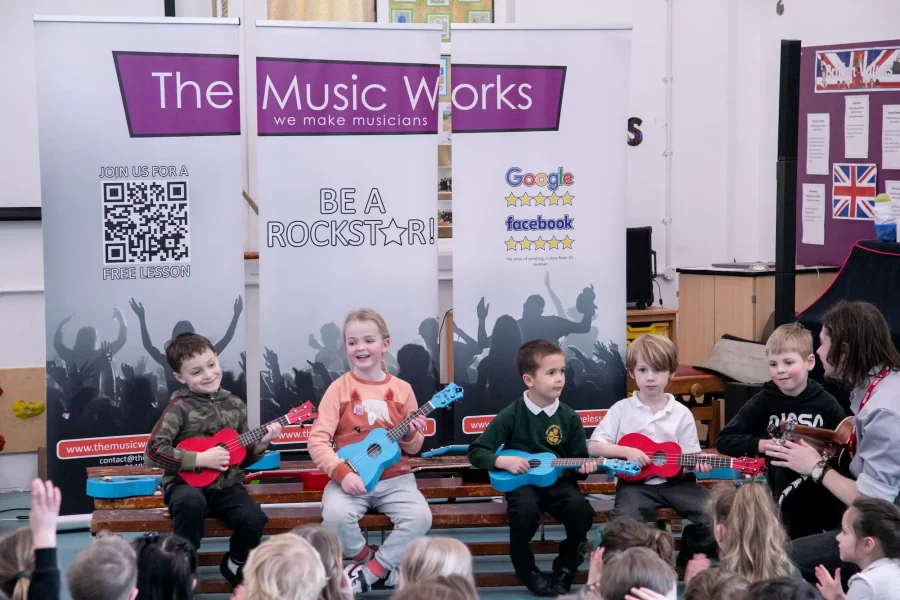 A group of children smiling and playing guitars during a school music workshop led by an instructor from The Music Works.