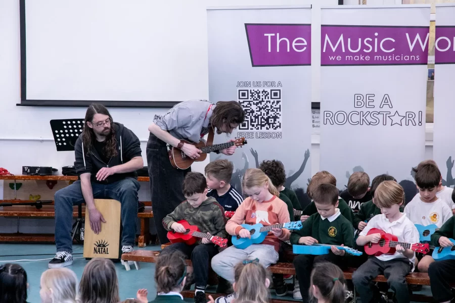 Children playing guitars while an instructor and a percussionist guide them during a school music workshop by The Music Works.