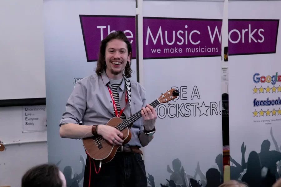 A smiling music instructor from The Music Works playing the ukulele during a school music workshop.