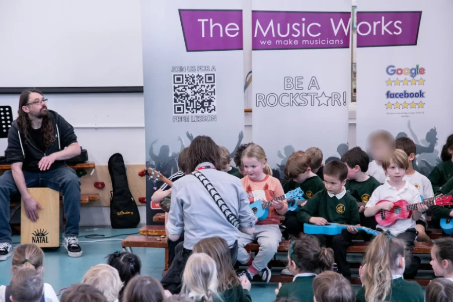 Children playing guitars while a percussionist accompanies them on a cajón during a school music workshop by The Music Works.