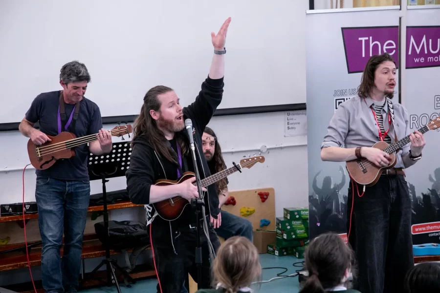 A group of musicians performing with ukuleles, a bass guitar, and percussion during a school music workshop by The Music Works.