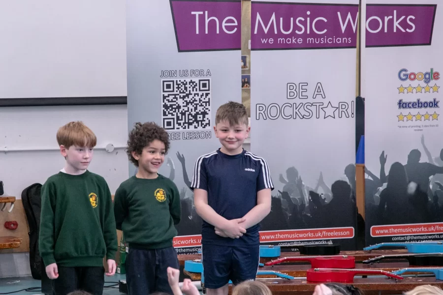 Three children stand proudly in front of The Music Works banners after participating in a school music workshop.