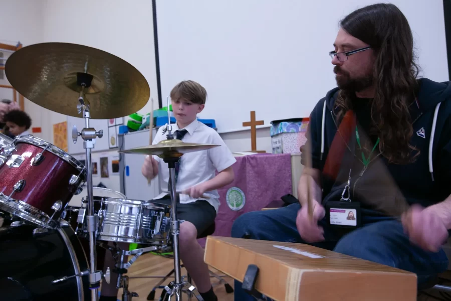 A boy in a white shirt playing drums while a music teacher plays a cajón beside him