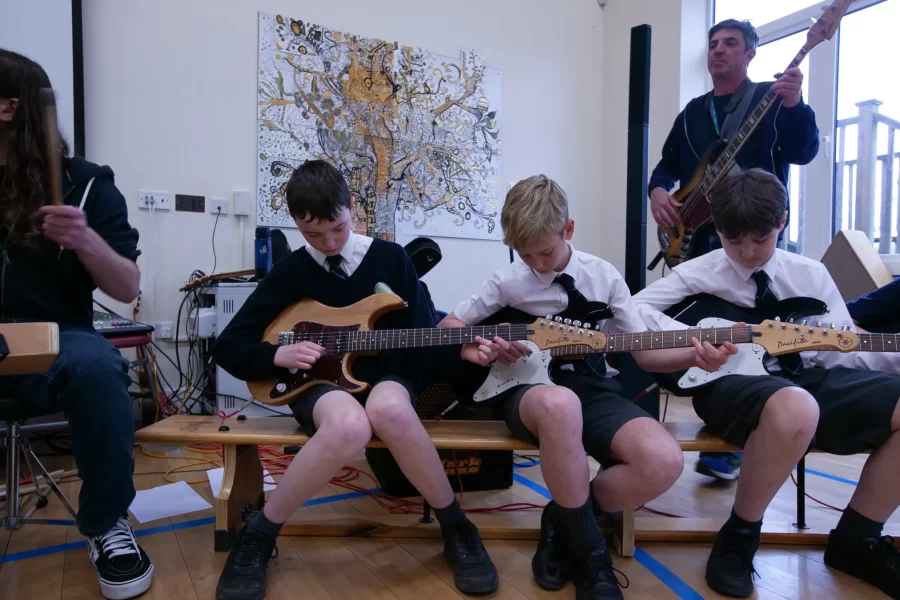Three boys in school uniforms playing electric guitars while a teacher plays bass behind them