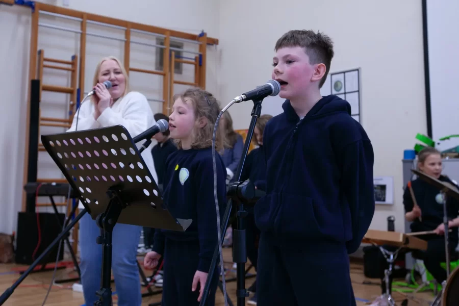 Two children singing into microphones on stage while a teacher sings behind them in a school performance.