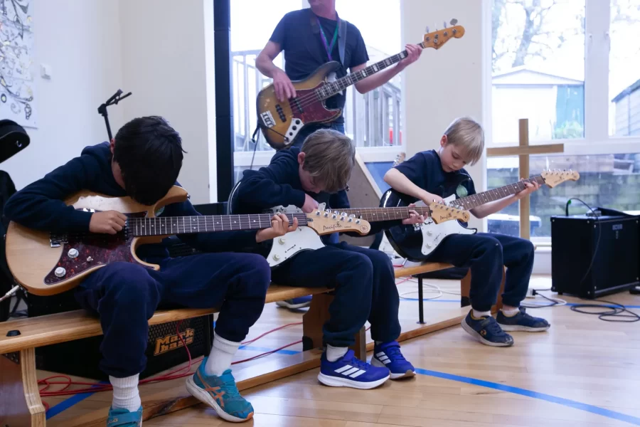Three young boys in navy uniforms play electric guitars while an instructor plays bass in the background