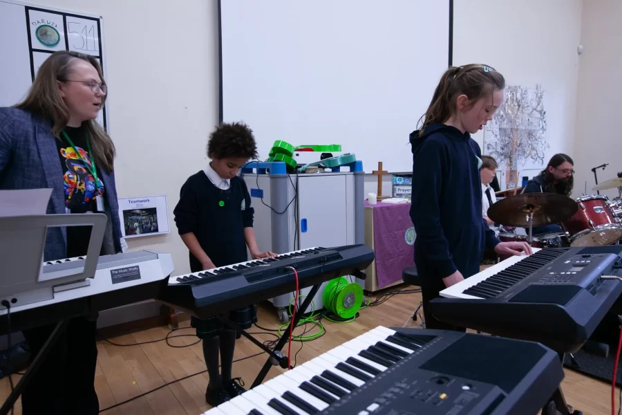 Two girls and a music teacher play keyboards during a school performance, with a drummer in the background.