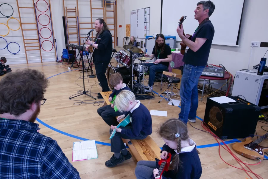 A group of students play guitars, ukuleles, and drums in a school music band performance led by teachers.