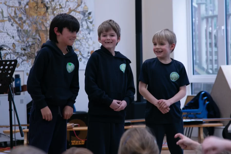 Three young boys in navy hoodies stand on stage smiling after a school music performance.