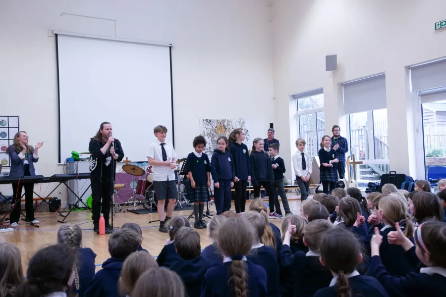 A group of students stand in front of an audience in a school hall, with a music teacher leading them on stage.