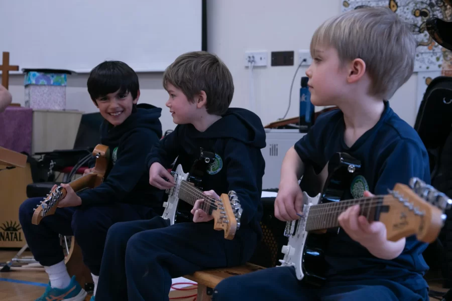 Three students in school uniforms sit on a bench, smiling as they play electric guitars.