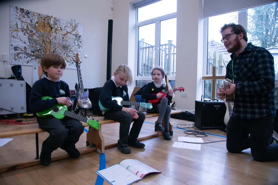 Three young students sit on a bench playing ukuleles while a teacher kneels beside them, offering guidance.