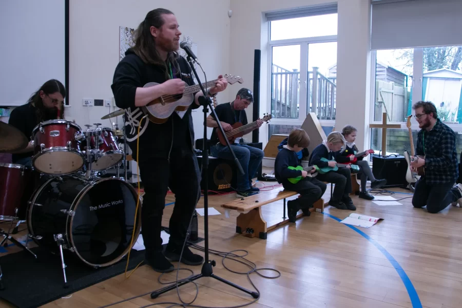 A teacher plays the ukulele while young students play ukuleles alongside a full school band.